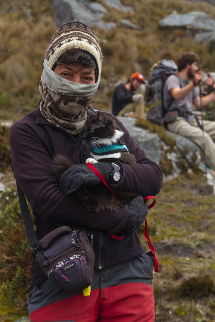 A person in winter wear holding a cat on a mountain hiking trail with others resting in the background.